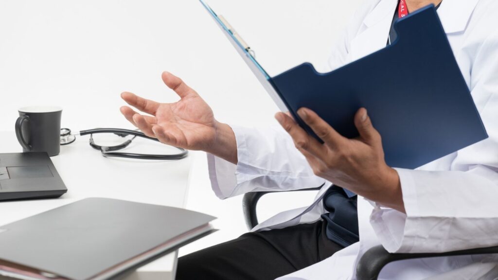 カルテを持って説明をする医師。
机にはパソコン、聴診器、カップがおいてある