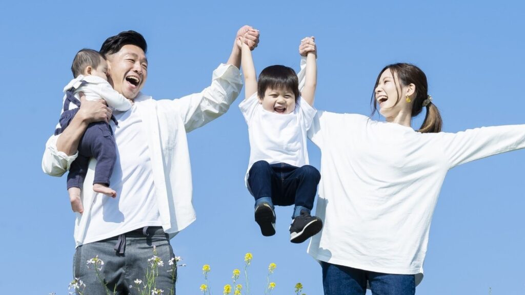 青空の下でママとパパに高い高いをしてもらっている男の子。パパは赤ちゃんを抱いている。